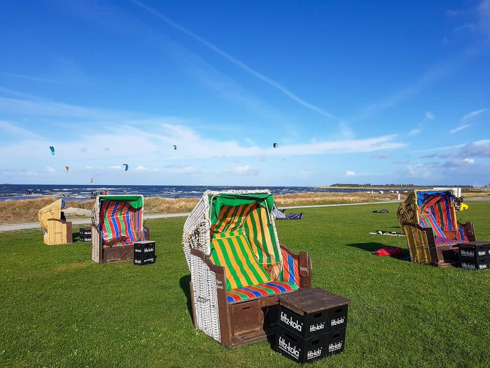 Colourful Strandkorb beach chairs on grass, with kites flying over the sea behind them.