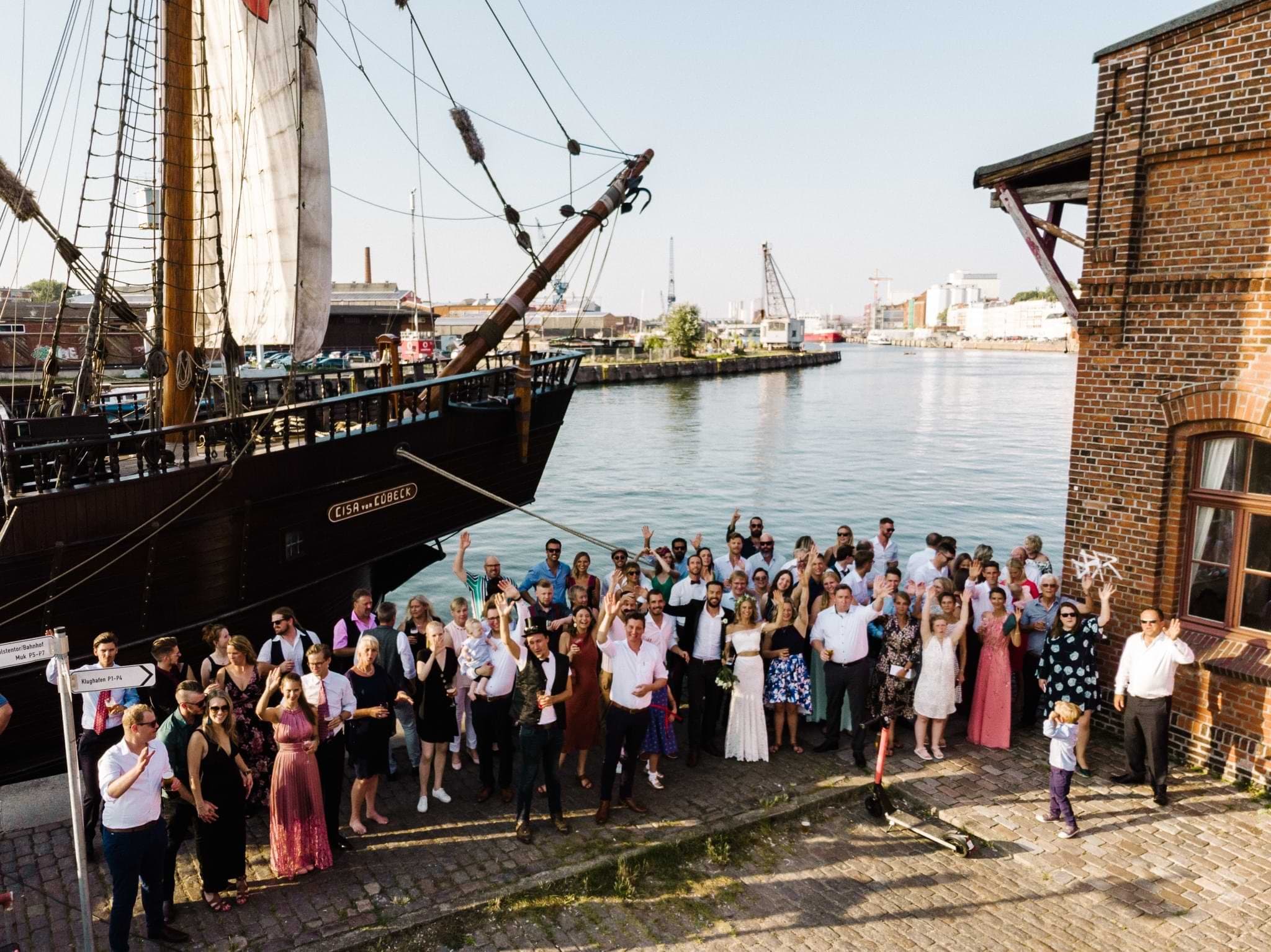 Wedding guests gathered on a harbour quay beside a tall sailing ship in Lübeck.