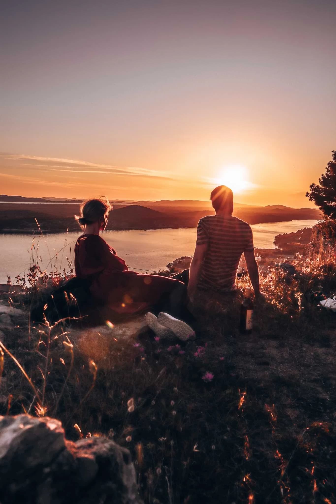 Two people sitting beside the water in Šibenik as the sun sets over distant hills.