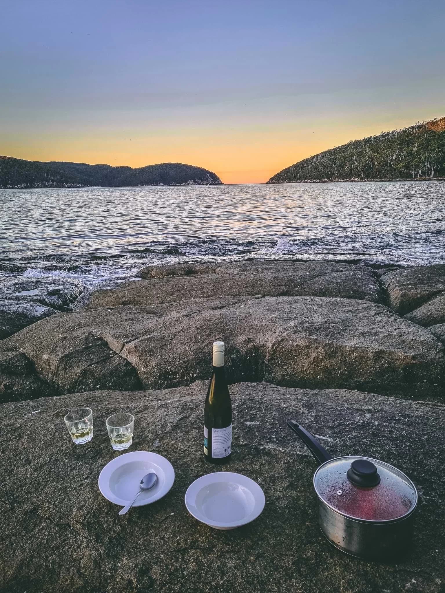 Camping cookware, plates and a bottle set on shoreline rocks at sunset.