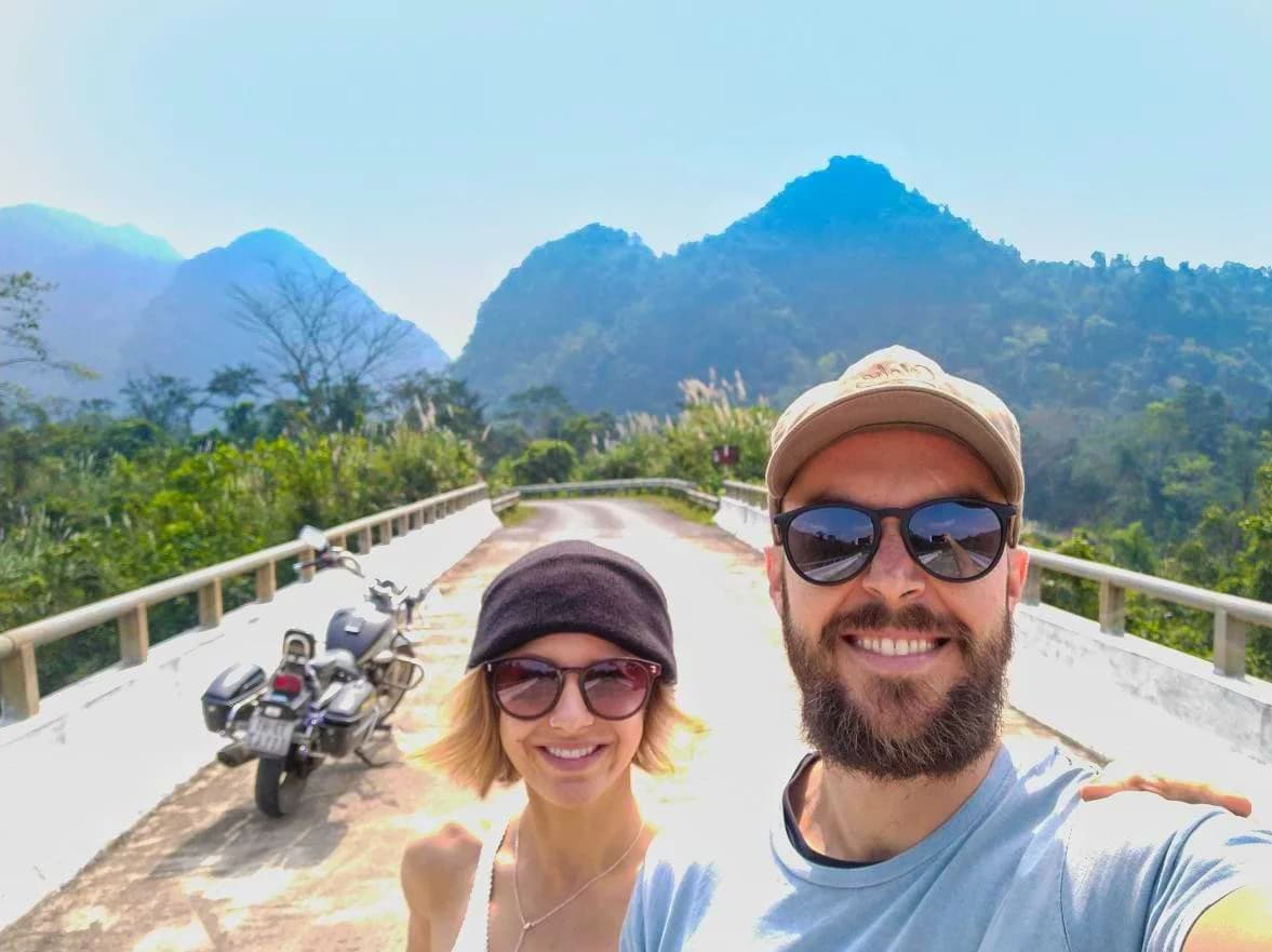 A couple taking a selfie beside their motorbike on a bridge surrounded by green mountains in Phong Nha, Vietnam.
