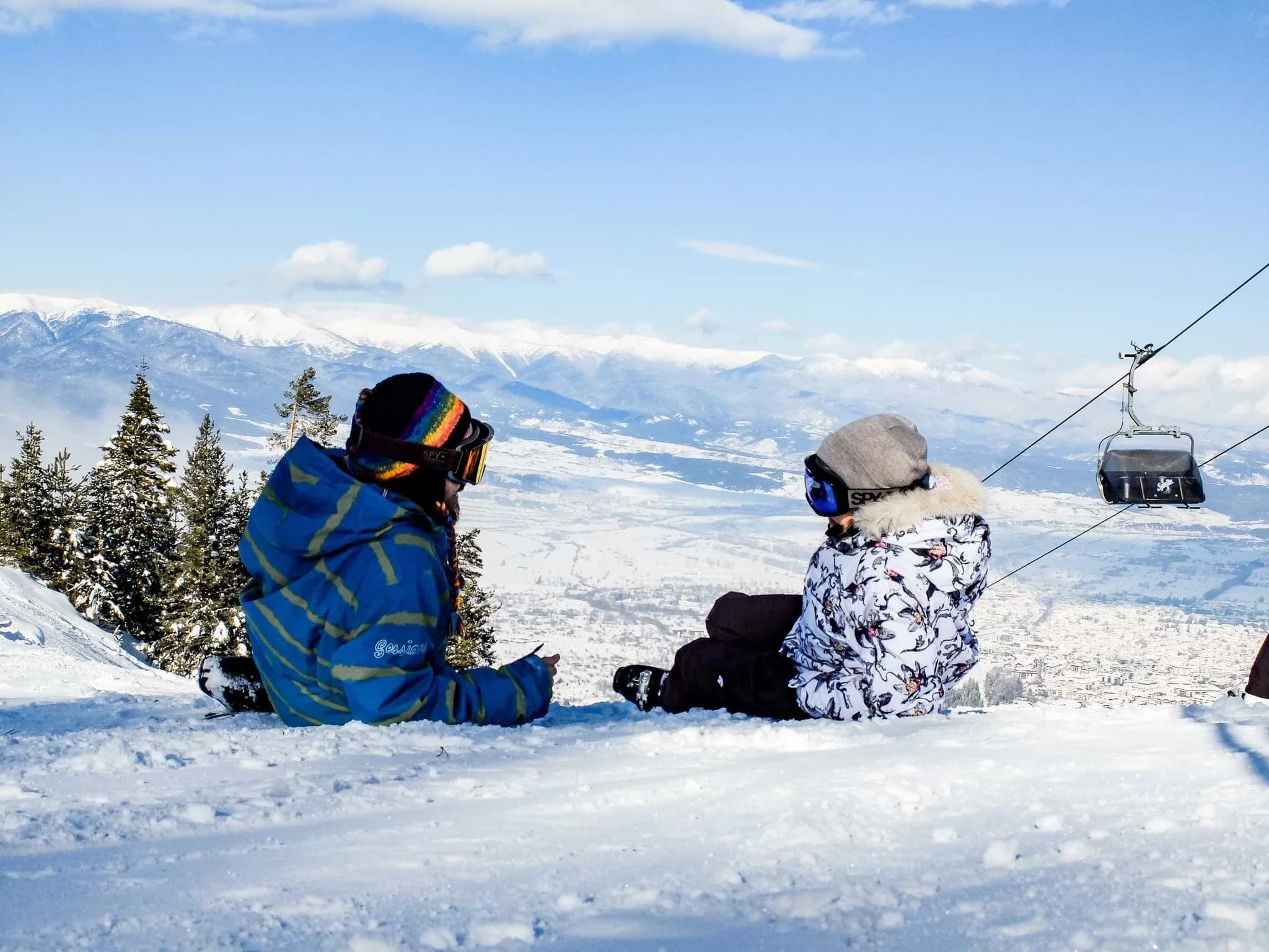 Two people resting on a snowy slope beside a gondola lift at Bansko ski resort.