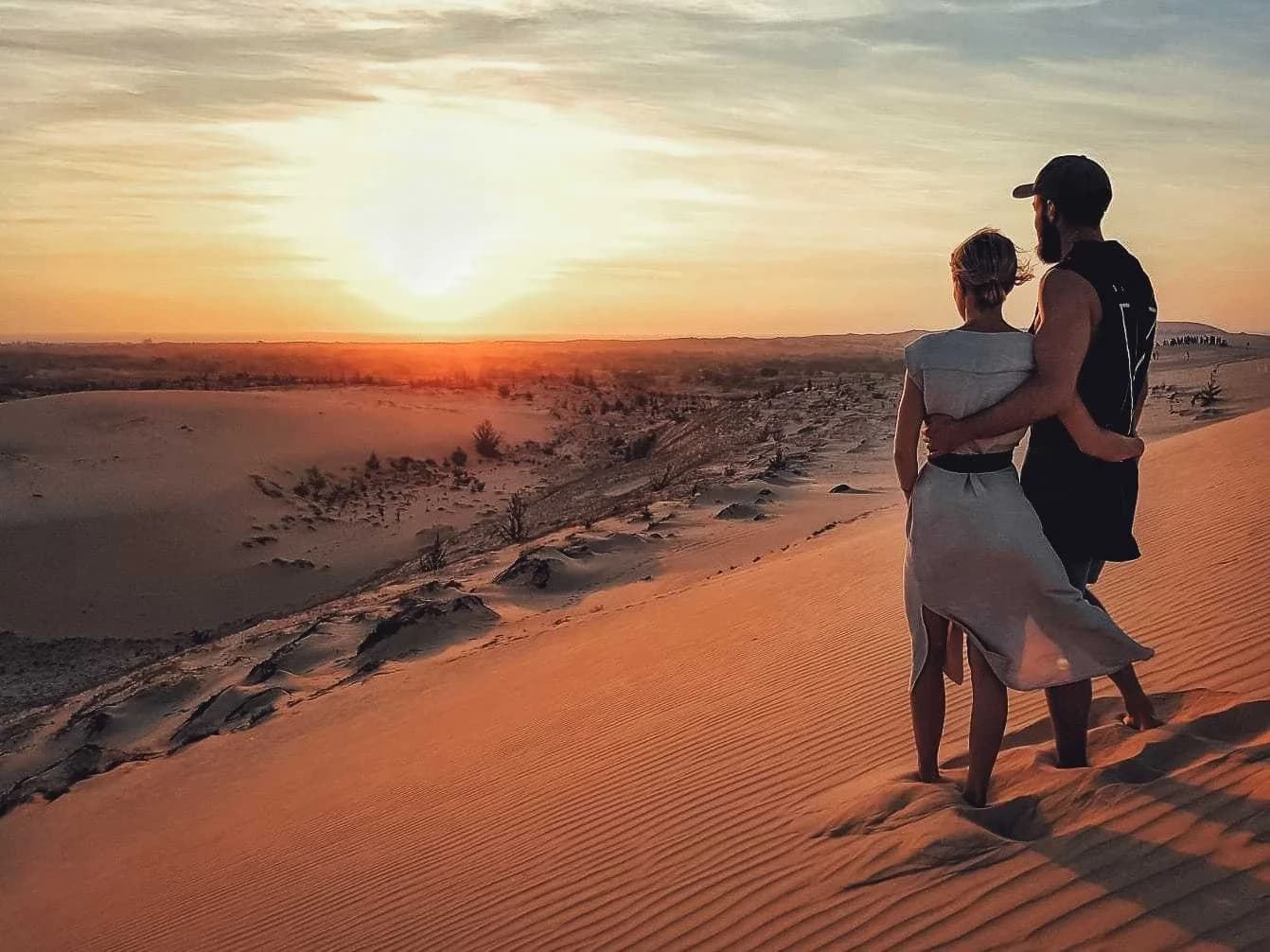 A couple standing together on sand dunes at sunset.