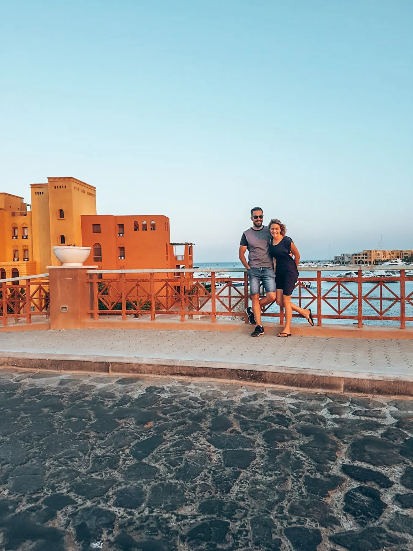 Two travellers standing on a bridge beside orange waterfront buildings and moored boats.