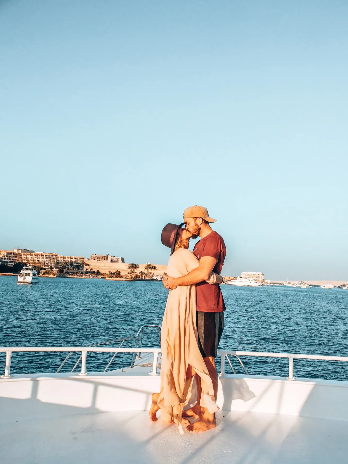 A couple standing on the bow of a boat with blue water and waterfront buildings behind them.