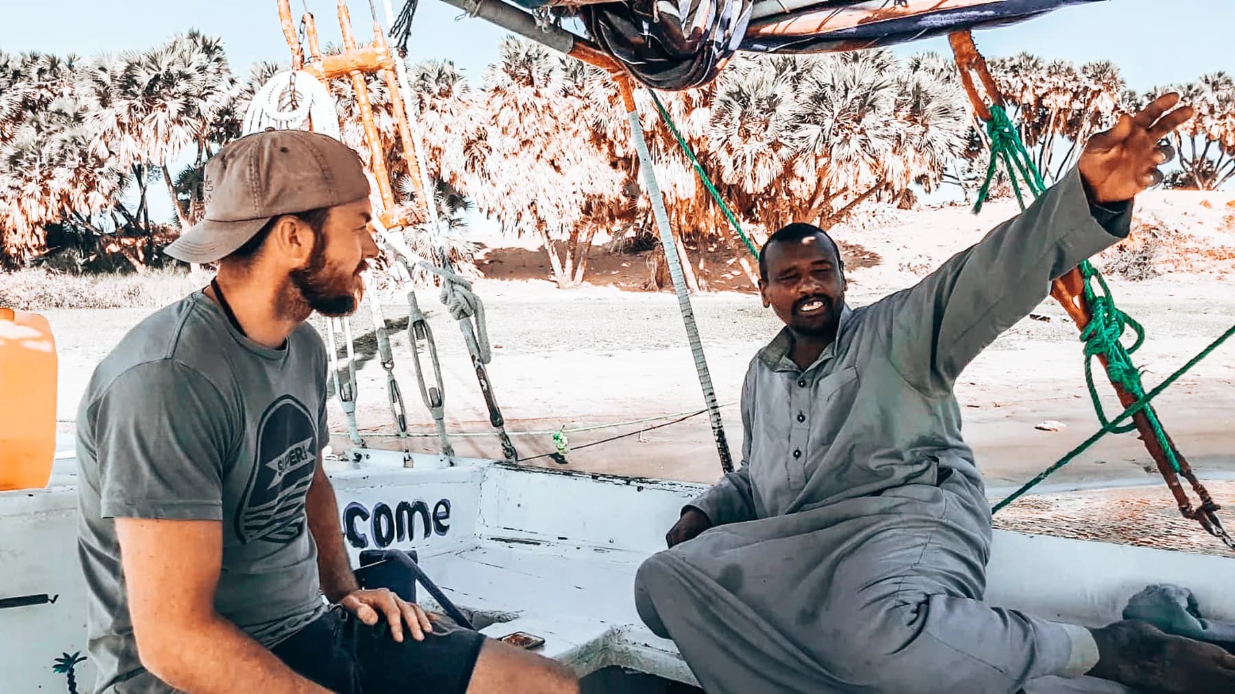 Two men sitting beneath a boat canopy beside a palm-lined riverbank.