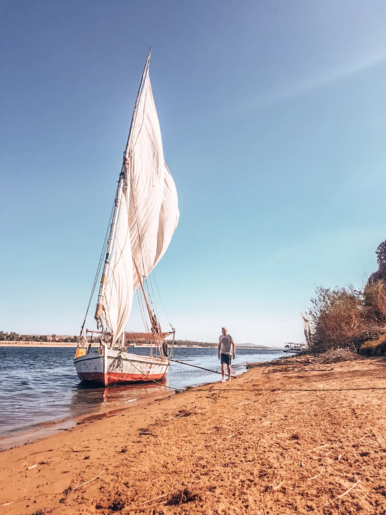 A traveller standing beside a moored sailboat on a sandy riverbank.