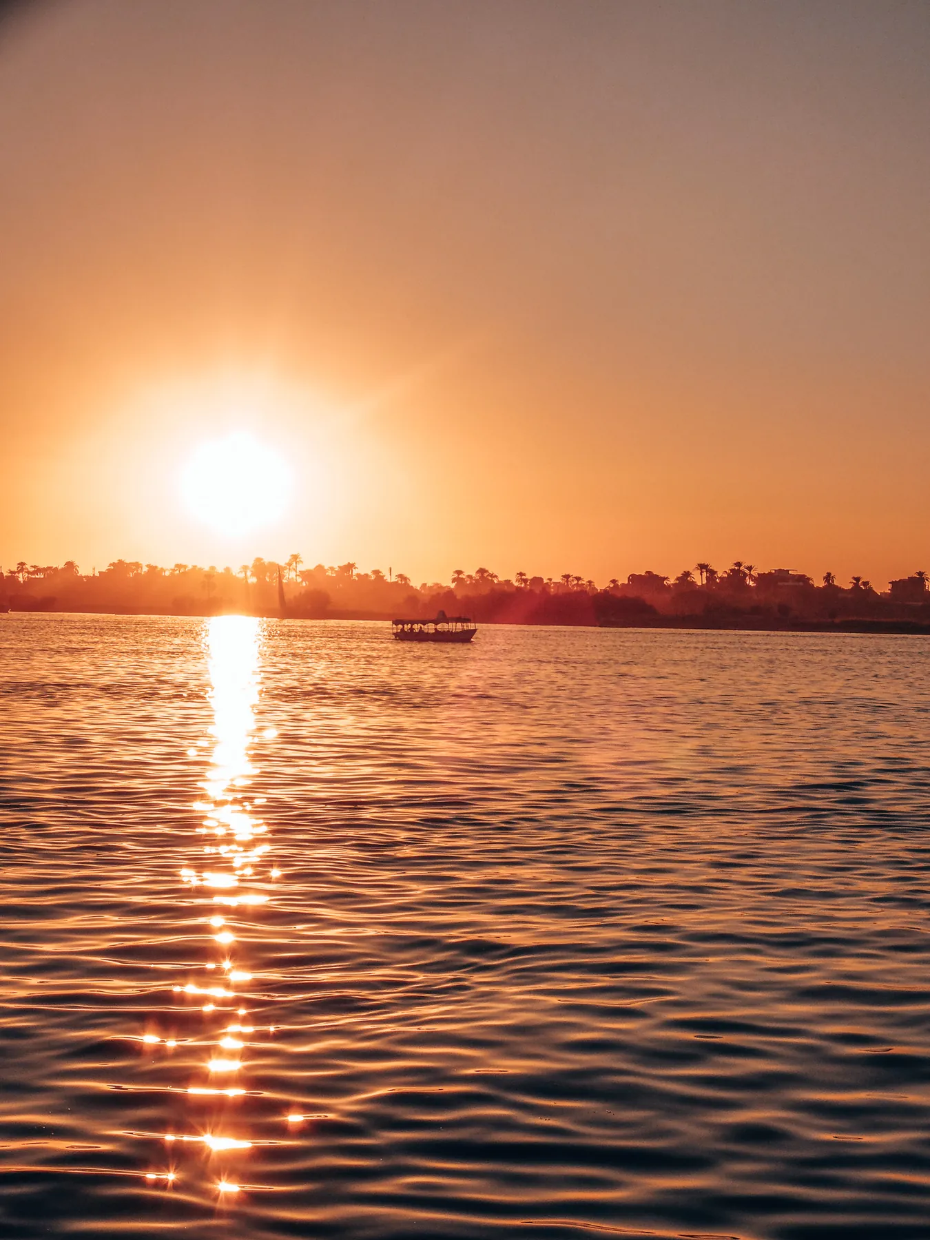 Sunset reflected on the Nile, with a small passenger boat and palm trees along the far bank.