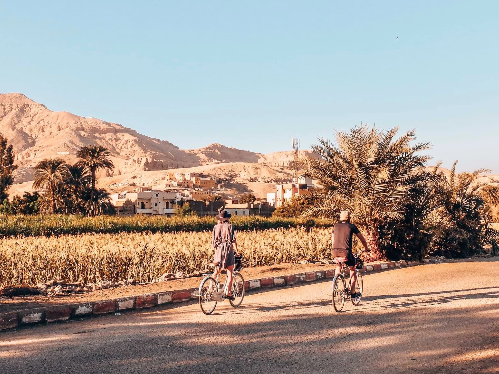 Two cyclists passing fields and palm trees beneath Luxor’s desert hills.