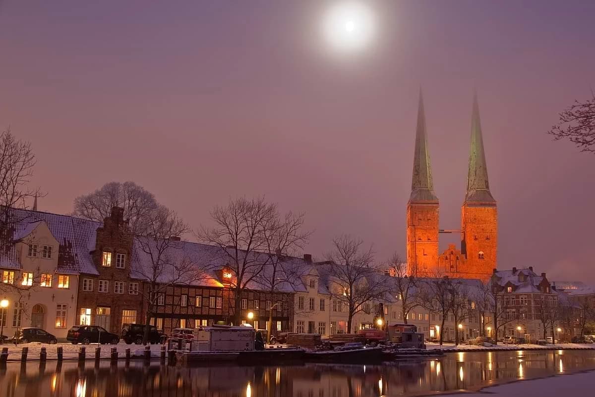 Snow-covered waterfront buildings and illuminated twin church spires beneath a bright moon.