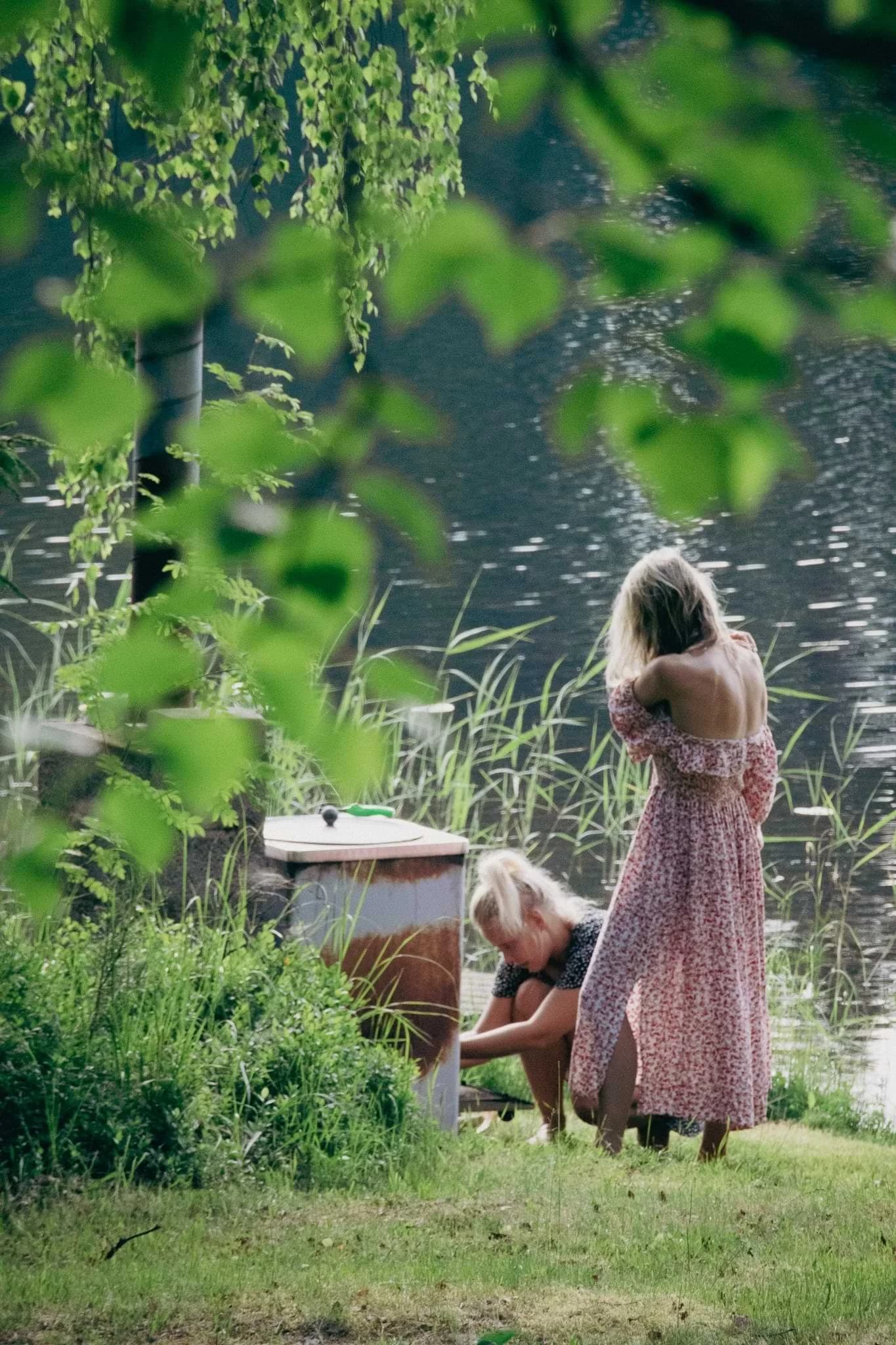 Two people beside a small outdoor table on a grassy riverbank shaded by trees in Finnish Lapland.