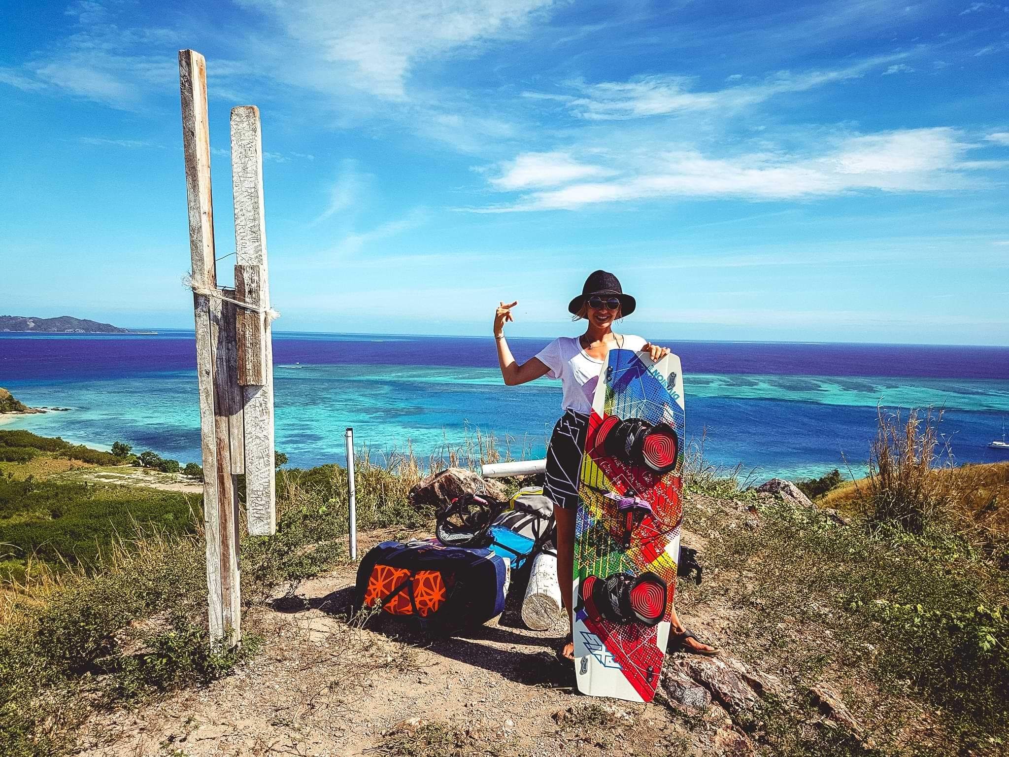 A person holding a kiteboard beside their equipment on a hillside overlooking a turquoise bay on Mana Island, Fiji.