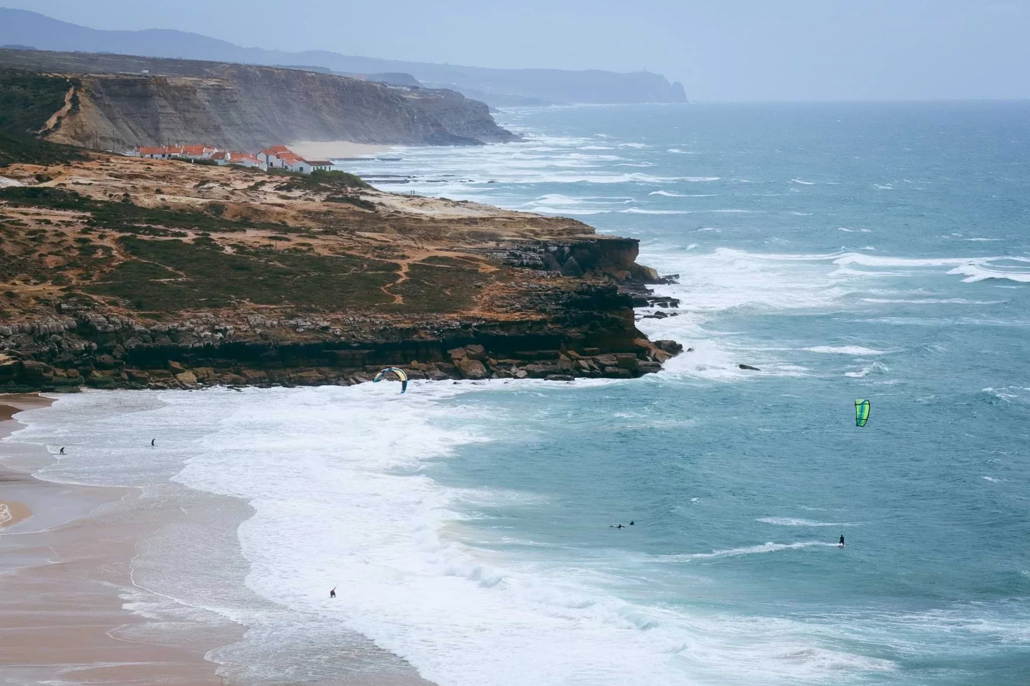 Kitesurfers riding waves beside a sandy beach and rugged coastal cliffs at Ericeira, Portugal.