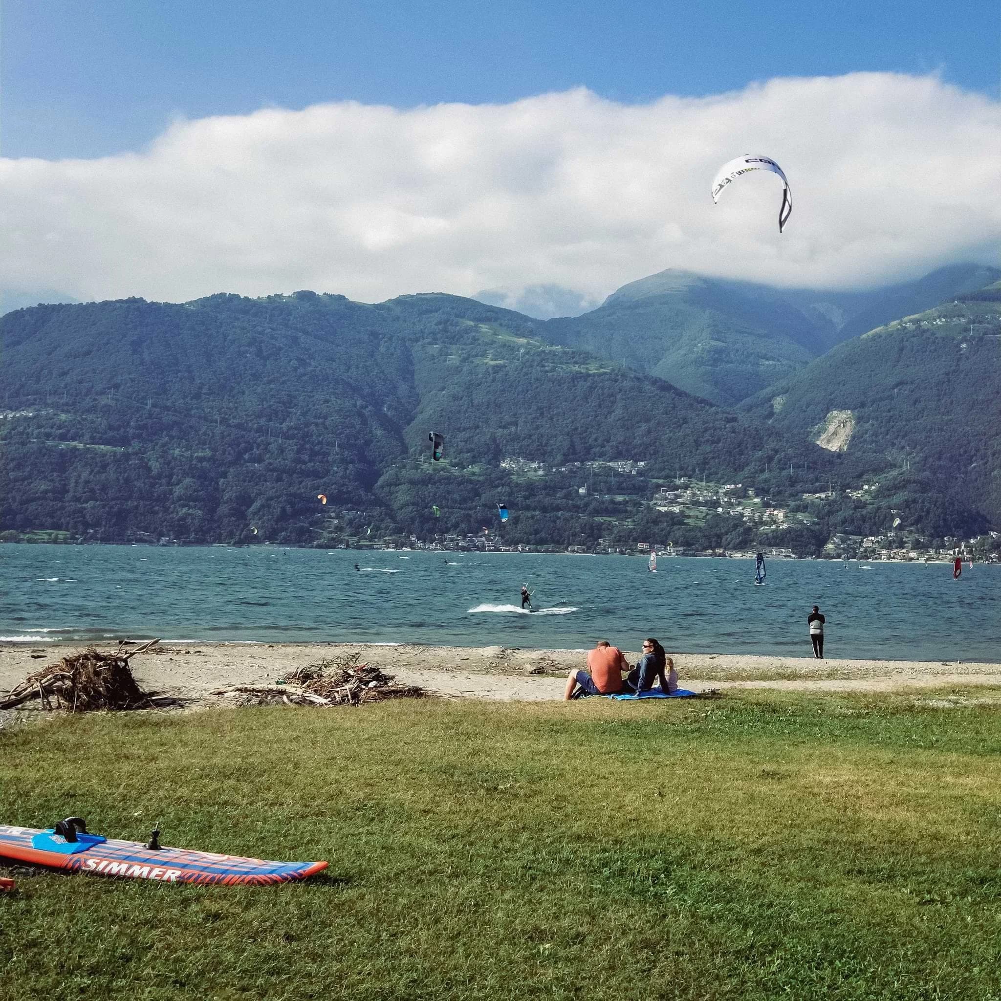 Kitesurfers on Lake Como beside a grassy shore, with wooded mountains behind them.