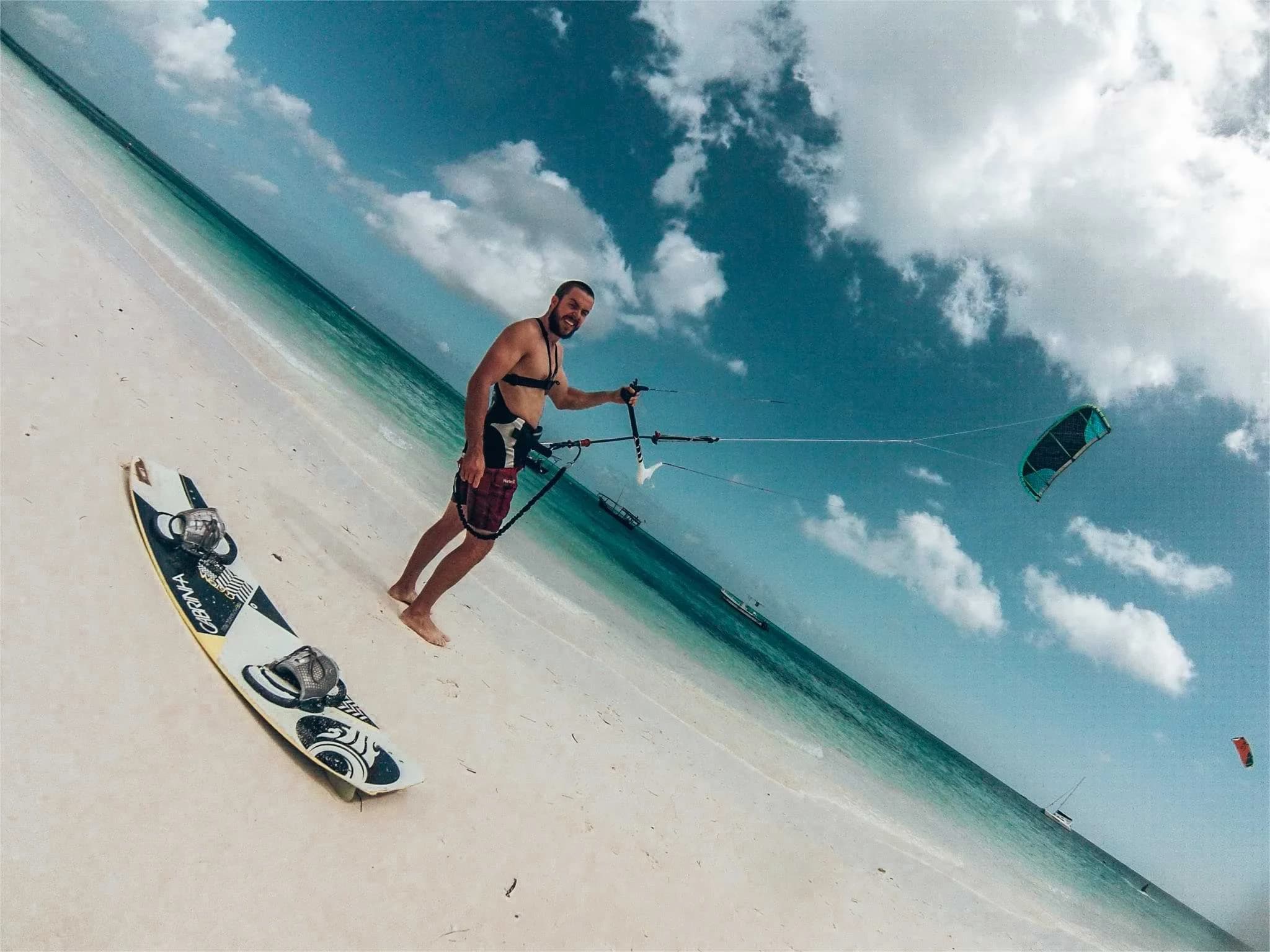 A kitesurfer standing on a white-sand beach in Zanzibar with a board and kite control bar.