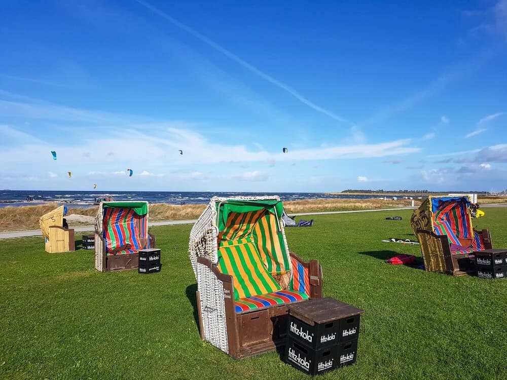 Colourful Strandkorb beach chairs on grass, with kites flying over the sea behind them.