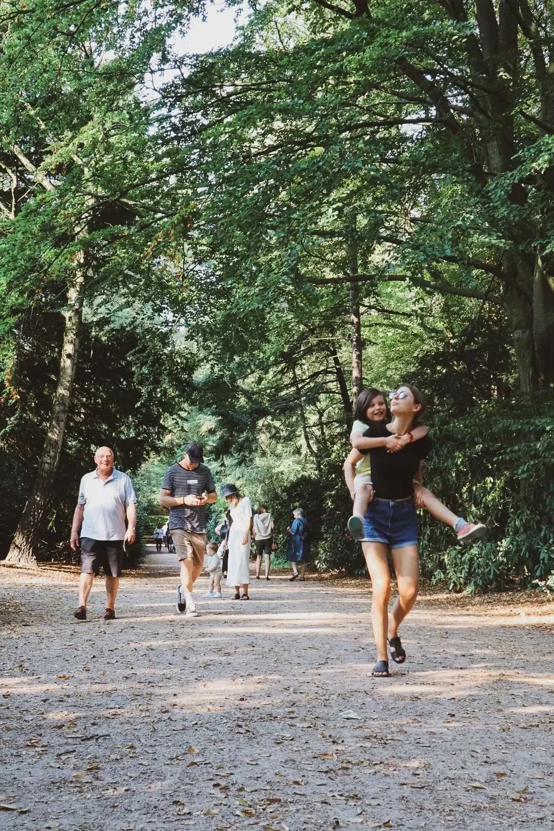 A person carrying another on their back along a tree-lined gravel path in Stadtpark, Hamburg, with other walkers nearby.