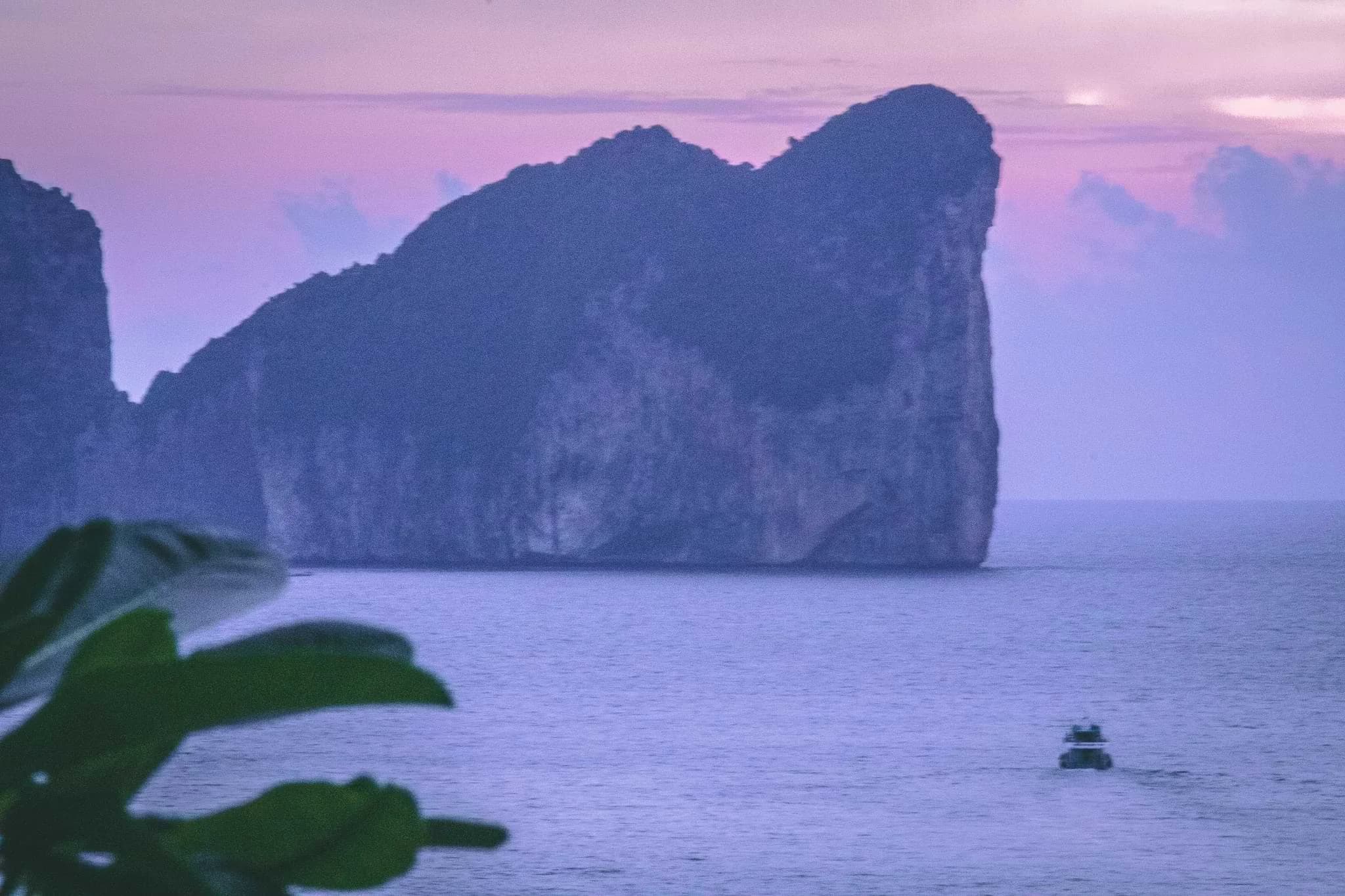 A small boat below a towering limestone island, viewed from Phi Phi Island in purple evening light.