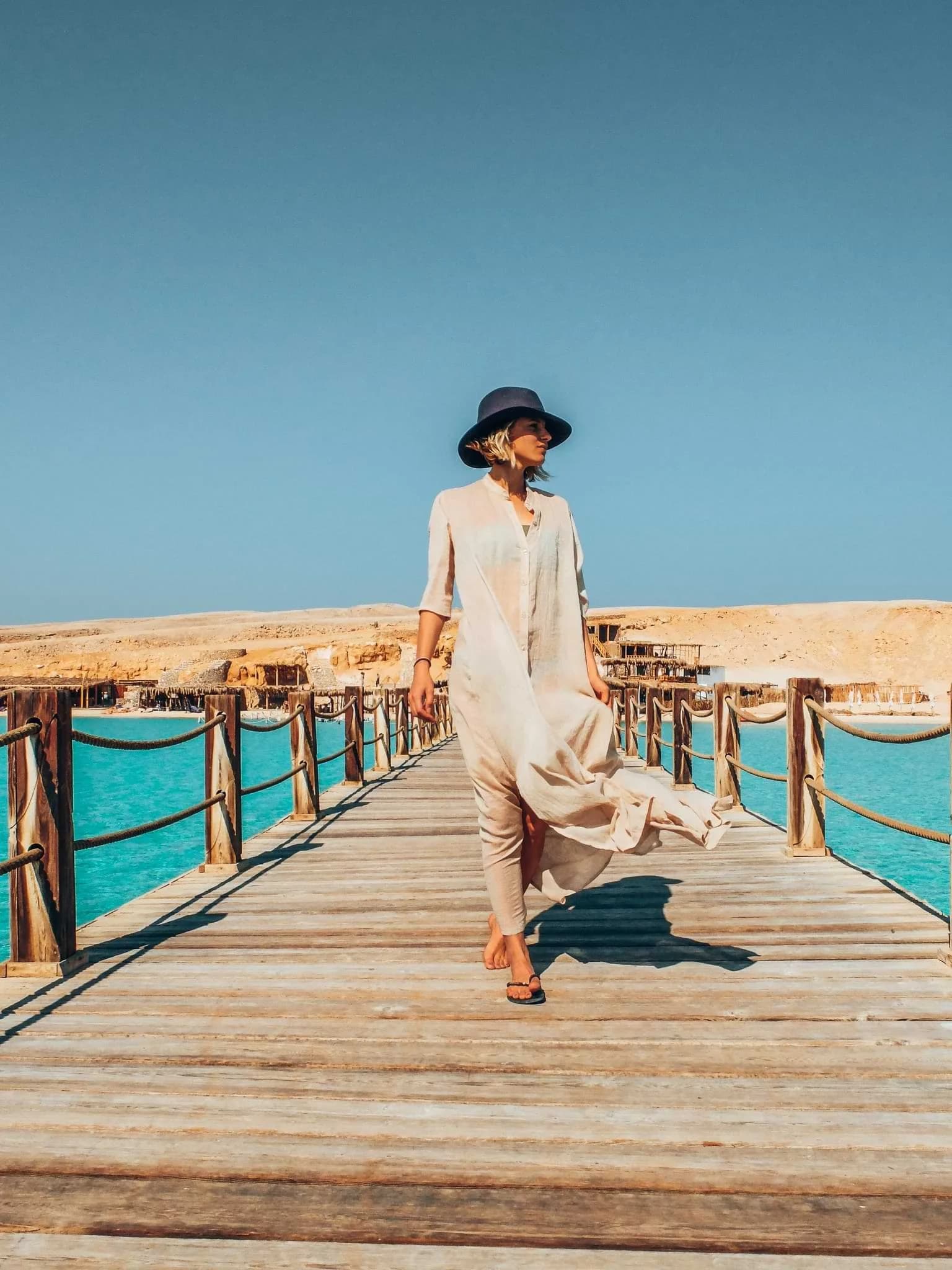 A person in a loose, light-coloured dress and hat walking along a wooden jetty over turquoise water.
