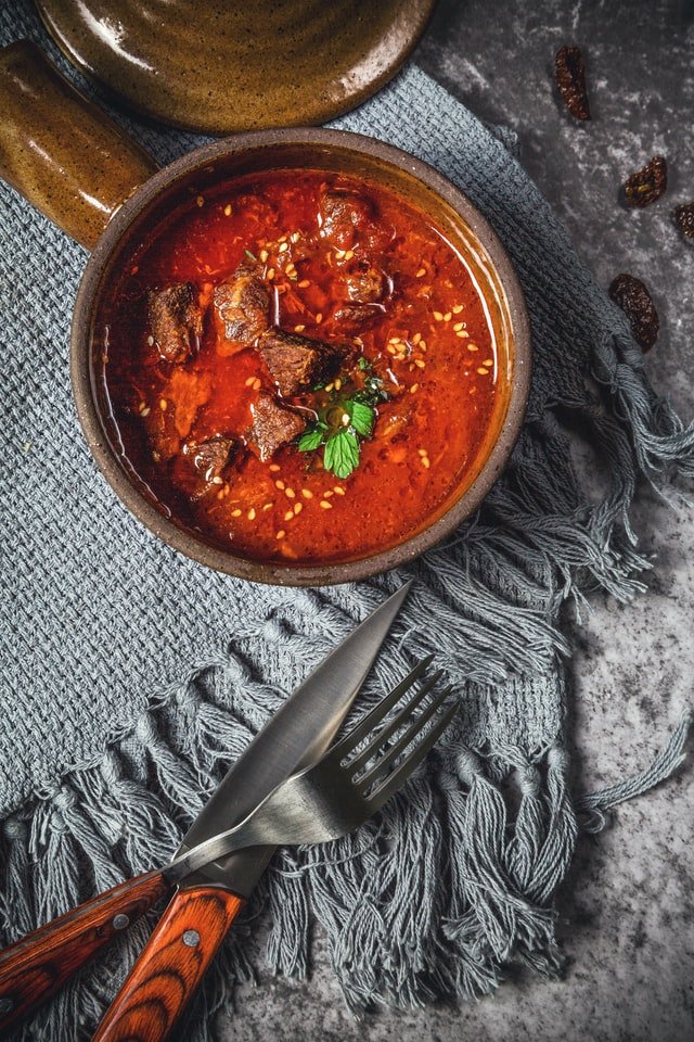 A bowl of red stew garnished with green herbs, beside cutlery on a textured cloth.