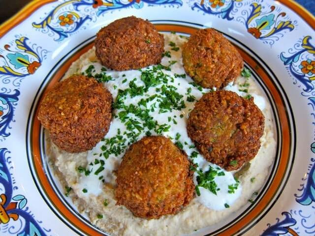 Five falafel pieces on a creamy dip, sprinkled with chopped herbs on a patterned plate.